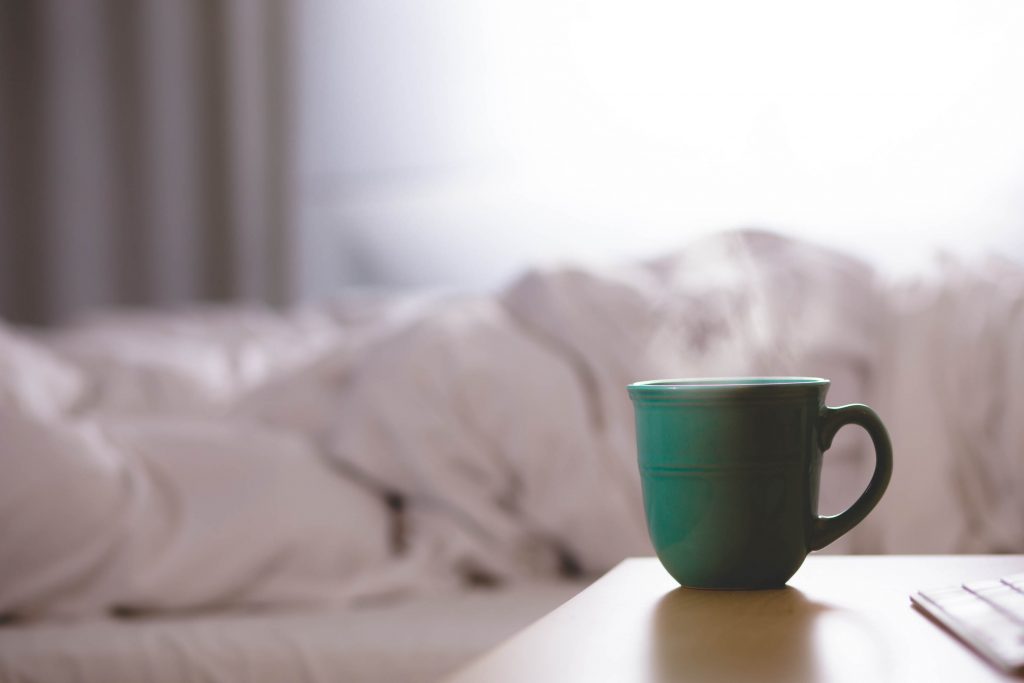 Mug of steaming drink on a bedside table with an unmade bed blurred in the back